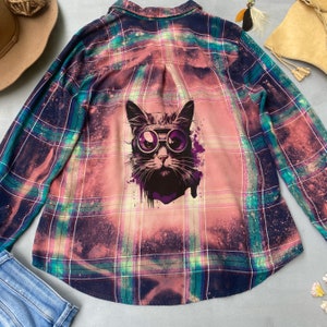 Puede incluir: Una camisa de franela a cuadros rosa, verde y negro con efecto blanqueado. La camisa presenta una impresión gráfica de un gato con gafas en la espalda.