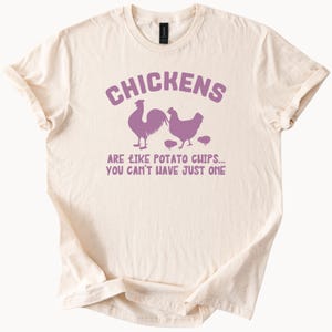 Puede incluir: Camiseta color crema con un gráfico morado de un gallo, una gallina y pollitos. El texto encima del gráfico dice "CHICKENS" y debajo dice "ARE LIKE POTATO CHIPS... YOU CAN'T HAVE JUST ONE."