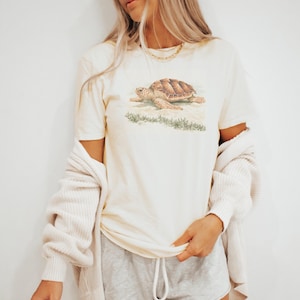 May include: A cream-colored short-sleeve t-shirt featuring a detailed illustration of a brown sea turtle on a sandy beach with green vegetation. The shirt is worn with a cream-colored ribbed cardigan and light gray drawstring shorts.