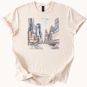 Puede incluir: Camiseta color crema con una ilustración en acuarela de Times Square en la ciudad de Nueva York. La obra de arte representa los edificios icónicos, las vallas publicitarias y los taxis. La camiseta tiene cuello redondo clásico y mangas cortas.