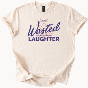 以下が含まれることがあります： ベージュのTシャツに、青と赤のグラフィックで「The Most Wasted of Days is One Without Laughter」と書かれています。