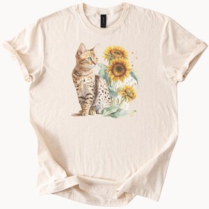 May include: A beige t-shirt featuring a brown and black tabby cat sitting in front of a sunflower. The cat is looking to the right and the sunflower is in bloom.