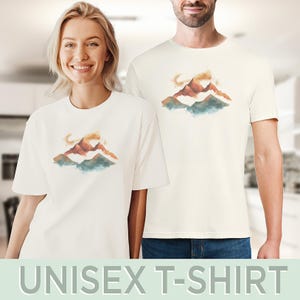 Op de afbeelding: Een wit unisex t-shirt met een aquarelontwerp van bergen in tinten oranje, bruin en blauw.