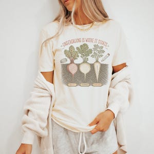 May include: A cream-colored short-sleeve t-shirt featuring a graphic of a beet, a turnip, and a parsnip growing in soil. The text reads "UNDERGROUND IS WHERE IT STARTS" at the top, with labels "BEET", "TURNIP", and "PARSNIP" below each vegetable illustration.