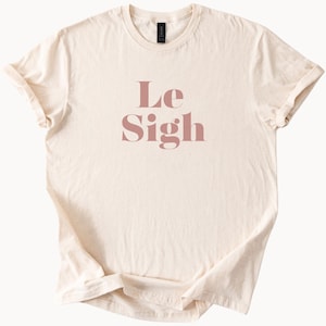 May include: Cream-colored t-shirt with the words "Le Sigh" printed in a dusty rose color. The shirt has short sleeves and a crew neck. The fabric appears soft and slightly wrinkled.