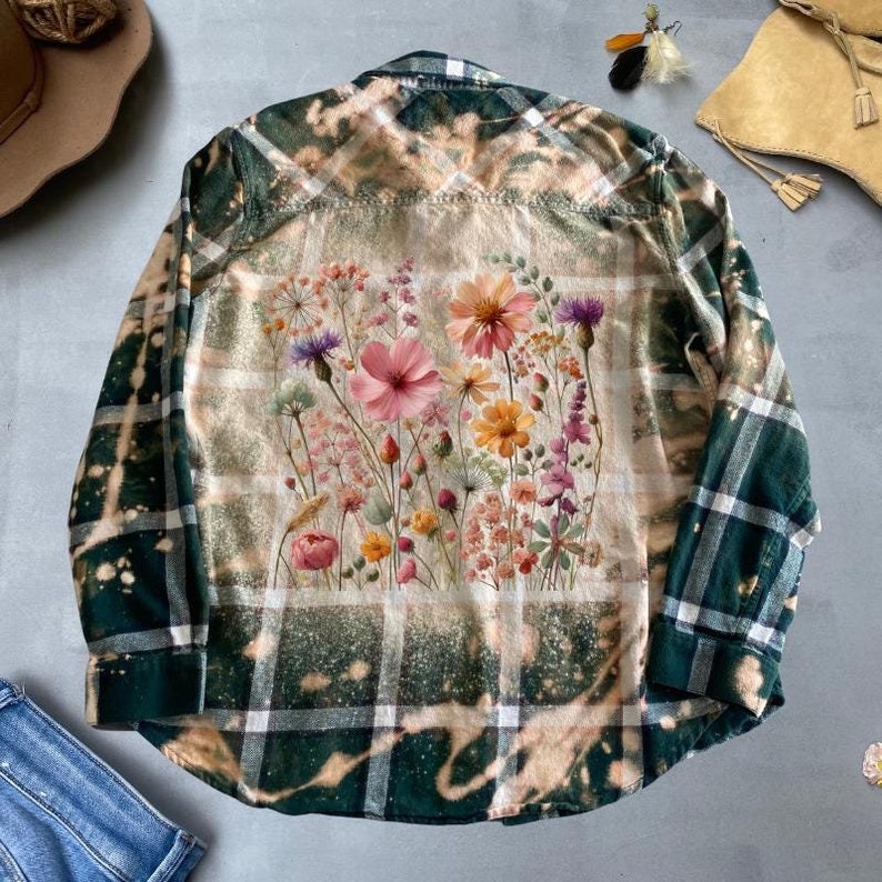 May include: A green and white plaid flannel shirt with a bleached effect and a floral print on the back. The floral print features a variety of flowers in shades of pink, yellow, and orange.