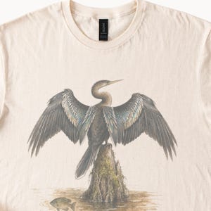 May include: A cream-colored t-shirt featuring a detailed graphic of a dark-feathered cormorant bird perched on a mossy tree stump in water, with its wings spread wide and a small fish swimming in the foreground below the stump.