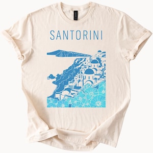 May include: A beige t-shirt with a blue graphic of the Santorini skyline. The graphic includes white buildings, blue flowers, and the text "SANTORINI".