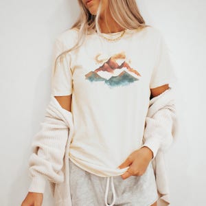 Op de afbeelding: Een wit T-shirt met een aquarelontwerp van bergen in tinten bruin, oranje en blauw.