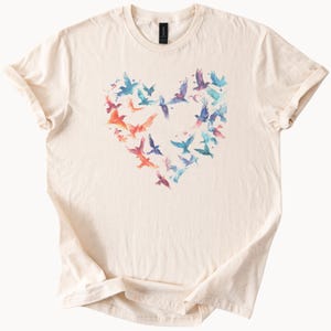 May include: Cream-colored t-shirt featuring a heart-shaped design composed of watercolor birds in shades of blue, orange, and purple. The birds are arranged to form a heart shape, creating a visually appealing and artistic design. The shirt has a classic crew neck and short sleeves.
