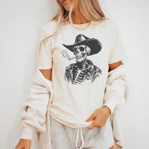 May include: Cream-coloured t-shirt featuring a black and white illustration of a skeleton wearing a cowboy hat and jacket, smoking a cigarette. The graphic is centered on the shirt.