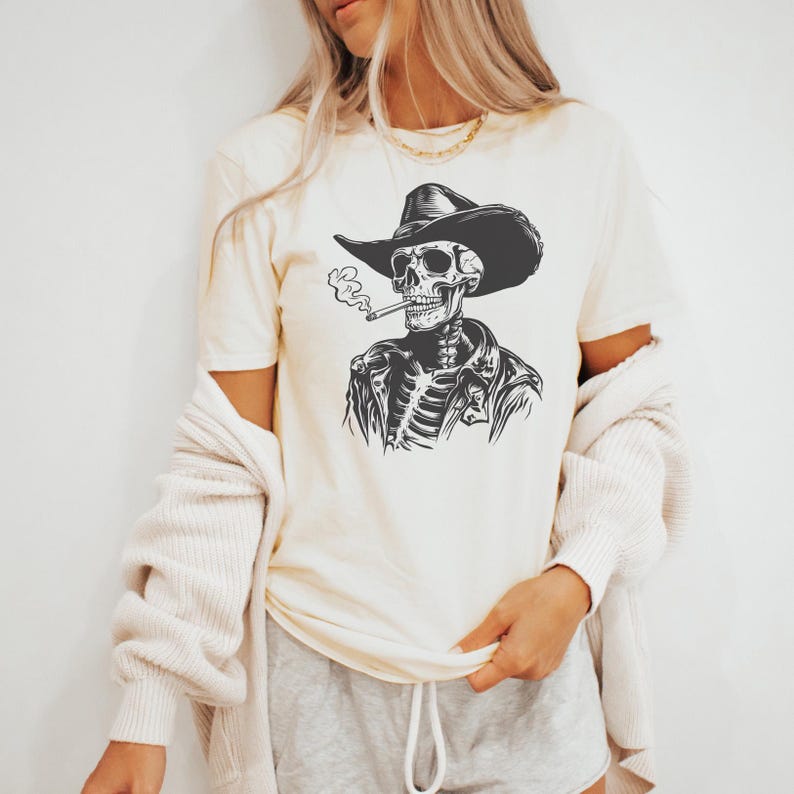 May include: Cream-coloured t-shirt featuring a black and white illustration of a skeleton wearing a cowboy hat and jacket, smoking a cigarette. The graphic is centered on the shirt.