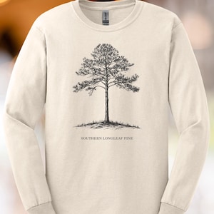 Puede incluir: Camiseta de manga larga color crema con una ilustración detallada en blanco y negro de un pino de hoja larga del sur. El texto "SOUTHERN LONGLEAF PINE" está impreso debajo del árbol. La camiseta tiene cuello redondo y mangas largas.