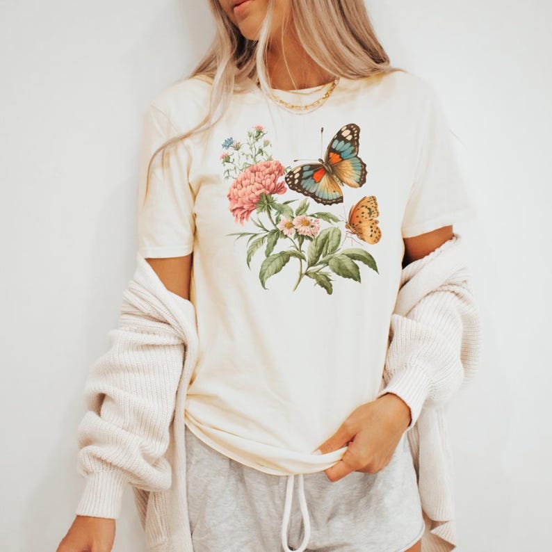 May include: Cream-colored t-shirt featuring a floral and butterfly design. The graphic includes pink flowers, green leaves, and two butterflies with orange, blue, and brown wings. The shirt has short sleeves.