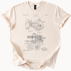 May include: Cream-colored t-shirt featuring a detailed black and white diagram of a tractor, including its internal components and design. The shirt has a crew neck and short sleeves. Text includes the date Sept. 26, 1933, and the name R. McCormick.