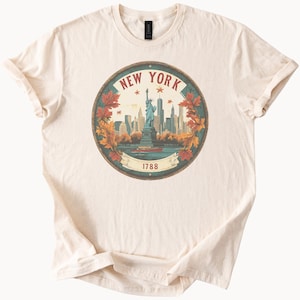May include: Cream-colored t-shirt featuring a circular graphic of the New York City skyline, the Statue of Liberty, and the text "NEW YORK 1788". The design includes fall foliage and stars, creating a vintage aesthetic.