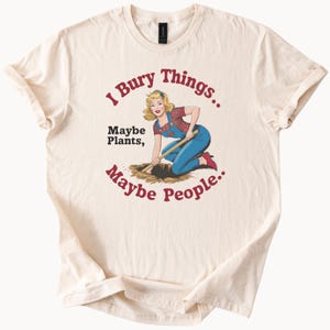 Puede incluir: Una camiseta beige con una ilustración de estilo vintage de una mujer cavando un agujero con una pala. El texto en la camiseta dice "I Bury Things... Maybe Plants, Maybe People."