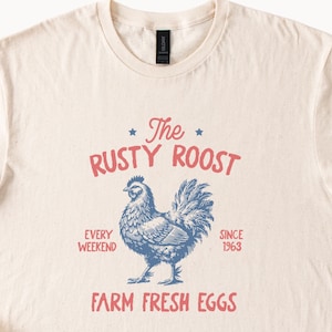 Op de afbeelding: Een beige T-shirt met een vintage-stijl grafisch ontwerp met een blauwe en witte illustratie van een hen. De tekst luidt "The Rusty Roost, Every Weekend, Since 1963, Farm Fresh Eggs".