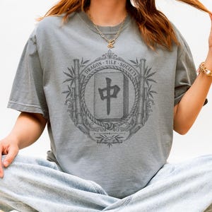 May include: A slate gray short-sleeve t-shirt featuring a dark gray graphic print of a mahjong tile with the Chinese character for middle, surrounded by a decorative circular frame of bamboo stalks, rope, and the text DRAGON TILE SOCIETY.