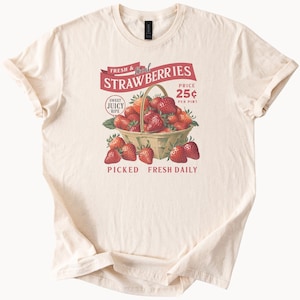 Könnte beinhalten: Ein weißes T-Shirt mit einer Vintage-Illustration eines Korbs voller roter Erdbeeren. Der Text lautet "FRESH & Local STRAWBERRIES", "SWEET JUICY RIPE", "PRICE 25¢ PER PINT", und "PICKED FRESH DAILY".
