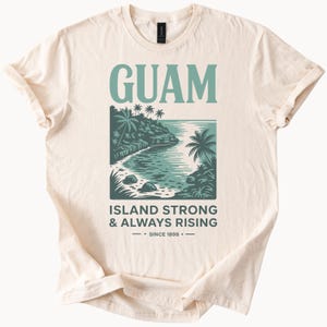 May include: Cream-coloured t-shirt with a teal graphic of a coastal scene, palm trees, and the word "GUAM". Below the image, the text reads "ISLAND STRONG & ALWAYS RISING - SINCE 1898".