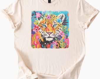 May include: A beige t-shirt with a colorful graphic print of a leopard's face surrounded by flowers. The leopard has blue eyes and a black nose. The flowers are pink, purple, yellow, and green.