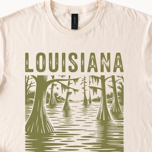 May include: Cream-colored t-shirt with the word "LOUISIANA" in green block letters. Below the text is a green illustration of a swamp scene with trees and water. The shirt has a crew neck and short sleeves.