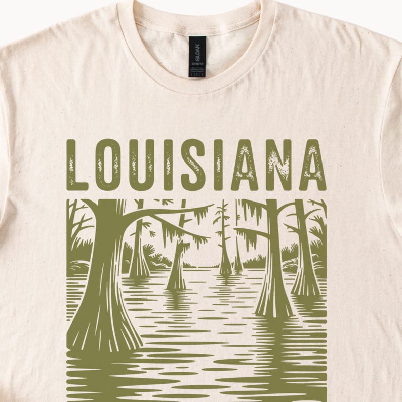 May include: Cream-colored t-shirt with the word "LOUISIANA" in green block letters. Below the text is a green illustration of a swamp scene with trees and water. The shirt has a crew neck and short sleeves.