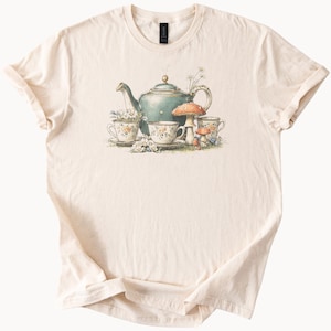 May include: Cream-colored t-shirt featuring a whimsical illustration. A teal teapot, floral teacups, and mushrooms are depicted. The design has a vintage, storybook aesthetic, with delicate floral details and soft color palette.