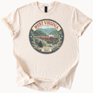 Puede incluir: Camiseta color crema con un diseño gráfico circular de West Virginia. El diseño incluye un tren cruzando un puente, un oso, montañas y el texto "WEST VIRGINIA" y "1863". La camiseta tiene mangas cortas y cuello redondo.