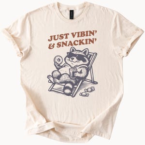 May include: Cream-colored t-shirt featuring a raccoon in sunglasses relaxing in a beach chair, holding a donut and a drink. The text "JUST VIBIN' & SNACKIN'" is printed above the raccoon in brown letters. The shirt is made of soft material.