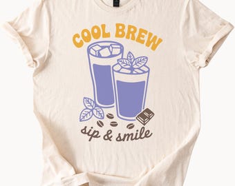 以下が含まれることがあります： クリーム色のTシャツ。氷、ミントの葉、コーヒー豆が入った2つのアイスコーヒーのグラフィック。テキスト「COOL BREW sip & smile」は黄色と茶色で印刷されています。