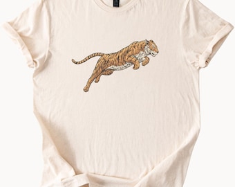May include: Cream-colored t-shirt featuring a detailed illustration of a leaping tiger. The tiger is depicted in shades of brown and orange with black stripes, showcasing a dynamic pose. The shirt is made of soft material.
