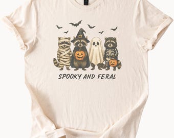 Op de afbeelding: Crèmekleurig T-shirt met een Halloween-thema. Het ontwerp toont vier wasberen verkleed als mummie, heks, spook en skelet, elk met een pompoen. De tekst "SPOOKY AND FERAL" staat eronder, met vleermuizen erboven.