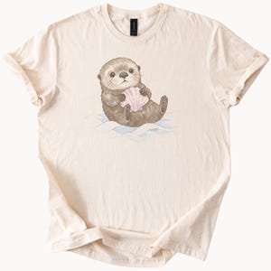 Op de afbeelding: Crèmekleurig T-shirt met een aquarel illustratie van een bruine otter die een roze schelp vasthoudt. De otter zit op blauwe watergolven. Het shirt is gemaakt van zacht materiaal.