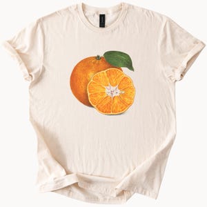 May include: Cream-colored t-shirt featuring a detailed illustration of an orange and a half-orange with a green leaf. The orange is a vibrant orange color, and the shirt has short sleeves.
