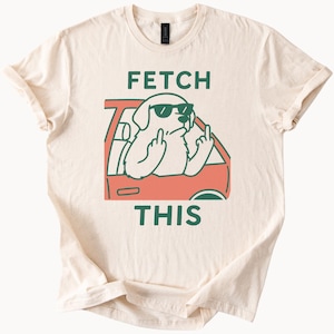 Może przedstawiać: Kremowa koszulka z kreskówkowym psem w okularach przeciwsłonecznych, pokazującym środkowy palec z czerwonego samochodu. Słowa "FETCH THIS" są wydrukowane na zielono nad i pod ilustracją psa.