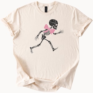May include: Cream-coloured t-shirt featuring a black skeleton graphic with a pink polka-dot bow tie. The skeleton is depicted in a running pose. The shirt has short sleeves and a crew neck.