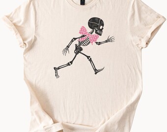 Puede incluir: Camiseta color crema con un gráfico de esqueleto negro con una pajarita rosa de lunares. El esqueleto está representado en una pose de carrera. La camiseta tiene mangas cortas y cuello redondo.