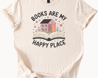 Może przedstawiać: Kremowa koszulka z napisem "BOOKS ARE MY HAPPY PLACE" w kolorze czarnym. Grafika przedstawia dom z różowymi drzwiami i oknem w kształcie serca, umieszczony na otwartej książce. Gwiazdy i kwiaty otaczają dom.