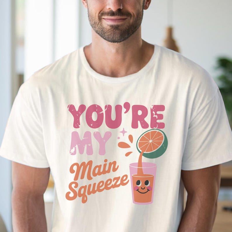 K&ouml;nnte beinhalten: Wei&szlig;es T-Shirt mit dem Text "YOU'RE MY Main Squeeze" in Pink und Orange. Eine Grafik einer halben Orange, die Saft in ein Glas mit einem l&auml;chelnden Gesicht gie&szlig;t, ist ebenfalls abgebildet. Das Shirt besteht aus einem weichen Material.