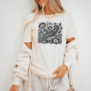 May include: A white t-shirt with a black and white graphic print of a bird in a floral design.