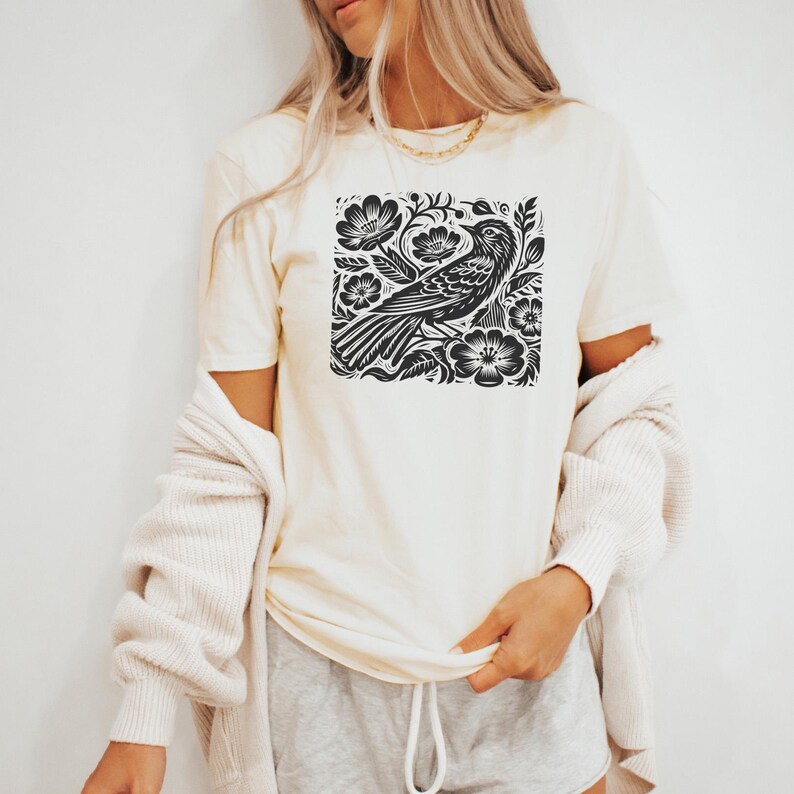 May include: A white t-shirt with a black and white graphic print of a bird in a floral design.