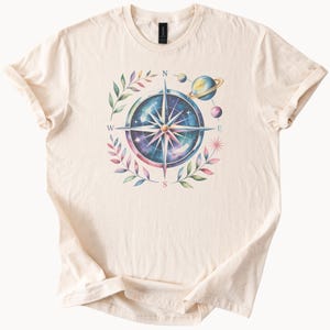 Op de afbeelding: Crèmekleurig T-shirt met een aquarelontwerp van een kompasroos, planeten en hemelse elementen. Het kompas is omgeven door bladeren en het shirt heeft opgerolde mouwen.