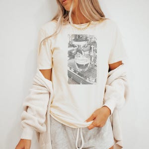 Puede incluir: Camiseta color crema con un boceto en blanco y negro de una máquina de espresso vertiendo café en un vaso transparente. La camiseta se combina con pantalones cortos grises y una rebeca color crema.