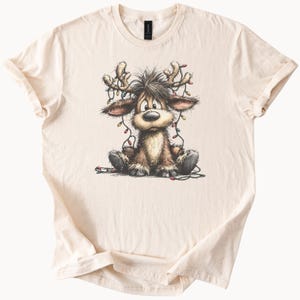 Puede incluir: Camiseta color crema con un reno de dibujos animados. El reno tiene pelaje marrón, cuernos adornados con luces navideñas de colores y una expresión ligeramente preocupada. El reno está sentado.