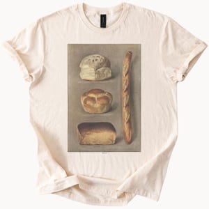 May include: Cream-colored t-shirt featuring a vintage illustration of various bread types: a round loaf, a brioche, a rectangular loaf, and a baguette. The word "BREAD" is printed at the bottom of the image.