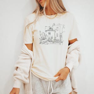 May include: Cream-colored t-shirt featuring a black line drawing of a building with plants and flowers. The shirt has short sleeves and a crew neck. A light beige cardigan is draped over the shoulders. The person is wearing gray shorts.