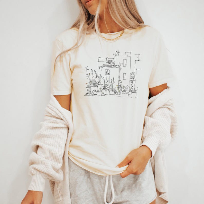 May include: Cream-colored t-shirt featuring a black line drawing of a building with plants and flowers. The shirt has short sleeves and a crew neck. A light beige cardigan is draped over the shoulders. The person is wearing gray shorts.