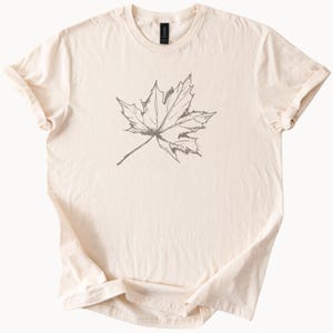 May include: A cream colored t-shirt with a simple line drawing of a maple leaf in gray.
